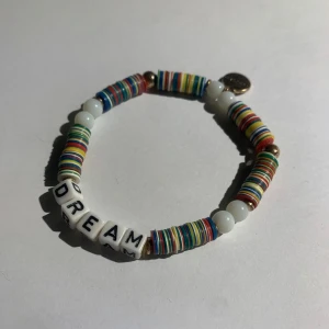 Färgglatt armband med bokstavspärlor - Säljer ett armband med färgglada platta pärlor och vita bokstavspärlor som bildar ordet 'DREAM'. Perfekt accessoar för att liva upp din stil!