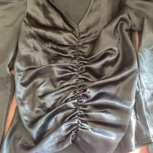 Svart glänsande topp med rynk  - Säljer en svart glänsande topp från Vero Moda i storlek XS. Topp har v-ringning, långa ärmar och snygga rynkdetaljer framtill som ger en smickrande passform. Materialet har en lätt glansig finish som känns lyxig.