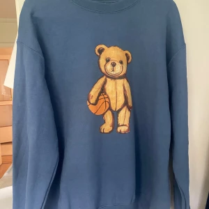 Blå sweatshirt med nalletryck  - Säljer en blå sweatshirt från Gildan med ett stort tryck av en nalle som håller i en basketboll på framsidan. Tröjan har rund hals och långa ärmar. Materialet är en blandning av bomull och polyester. Super skön och passar både tjejer och killar. Priset går att diskutera💞