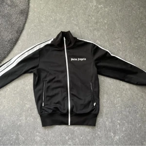 Svart track jacket från Palm Angels - Säljer en svart track jacket från Palm Angels med vita ränder längs ärmarna och vit dragkedja framtill. Jackan har hög krage och två fickor med dragkedja. Klassisk logga på bröstet. Den är äkta, köpt på kicksnstuff (kvitto finns) 