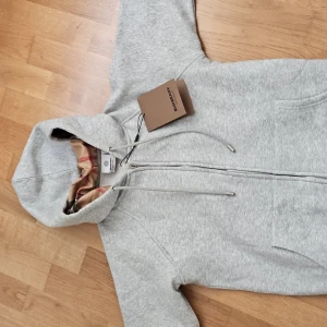Grå hoodie från Burberry med dragkedja - Pris kan diskuteras kan fixa sköna priser. Snygg ljusgrå hoodie från Burberry med klassiskt rutigt foder i huvan. Tröjan har dragkedja framtill, snörning i huvan och två fickor. Perfekt för dig som gillar stilrena och exklusiva plagg med en twist av ikoniskt mönster.
