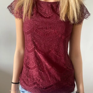 Vinröd spetsblus med kort ärm - Supersnygg vinröd blus i spets med korta ärmar och rund hals. Blusen har ett detaljerat spetsmönster över hela plagget och är lätt transparent på ärmarna. 