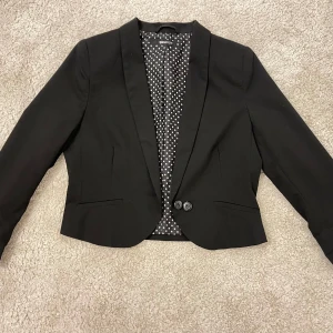 Svart kavaj Gina Tricot - Svart kavaj från Gina Tricot med prickigt foder. Kavajen har en klassisk krage och knäpps med två knappar framtill. I nytt skick
