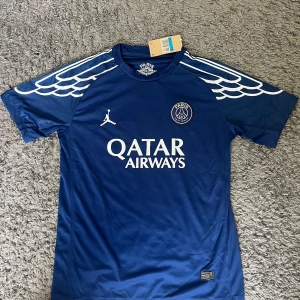 PSG x Jordan blå matchtröja - Storlek M. Blå PSG x Jordan fotbollströja med vita detaljer och vingar på axlarna. Korta ärmar och tryckt Qatar Airways-logga på bröstet. Tillverkad i lätt och ventilerande polyester, perfekt för träning eller match. Priset kan diskuteras.
