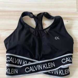 Svart sporttopp Calvin Klein - Sportig svart träningsstopp från Calvin Klein med bred resår och logga i vitt runt midjan. Toppen har meshdetaljer i ryggen och klassisk racerback-modell. Perfekt för träning och ger en snygg, tight passform. Har även padding vid brösten. 