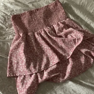 Rosa blommig volangkjol - Supersöt rosa kortkjol med vita små blommor och bred smock i midjan. Kjolen har volangdetaljer och omlottkänsla som ger en romantisk vibe. Perfekt för varma dagar när du vill känna dig extra gullig.