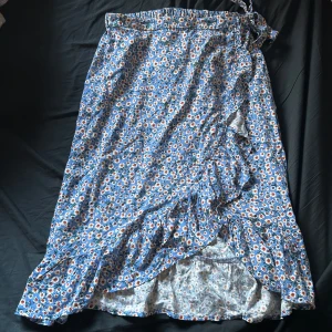 Blommig omlottkjol från JDY - Supersöt blå omlottkjol från JDY med blommigt mönster i vitt, gult och rött. Kjolen har volangdetaljer längs kanten och knyts i midjan. Perfekt för dig som gillar en feminin och somrig stil.