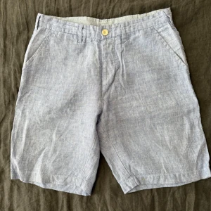 Ljusgrå linneshorts  - Snygga ljusgrå shorts  i svalt linnematerial. Klassisk rak modell med fickor fram och bak, bälteshällor och stängning med knapp. Perfekta för varma dagar när du vill ha en clean och enkel look.