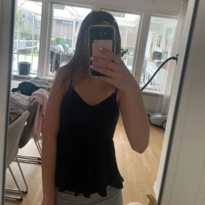 Svart ribbad volanglinne - Säljer ett svart kort linne med smala axelband och v-ringning. Enkel och stilren modell som sitter snyggt och är perfekt för varma dagar. Volangen  har en lätt utställd passform och är ärmlös för en luftig känsla.