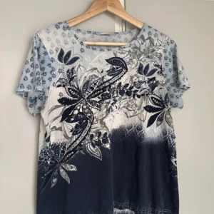 Blåvit blommig t-shirt i bomull - Snygg t-shirt i 100% bomull med blå och vita nyanser. T-shirten har korta ärmar och ett stort blommigt mönster framtill, samt små blommor över hela plagget. Perfekt för dig som gillar unika prints och mjuka material.