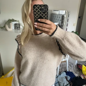Beige stickad tröja med volang Selected Femme - Superfin beige stickad tröja från Selected Femme med svarta kontrastdetaljer och volang längs axlarna. Väldigt bra skick då den inte har kommit till användning! Normal i storleken 