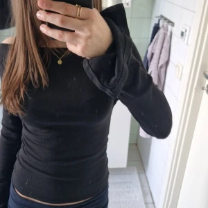 Svart offshoulder långärmad tröja - Svart långärmad offshoulder-tröja i tight passform. Tröjan är enkel och stilren med raka linjer och sitter snyggt mot kroppen. Perfekt att matcha med jeans eller kjol för en clean look. Materialet känns mjukt och stretchigt, troligen bomull eller polyester.