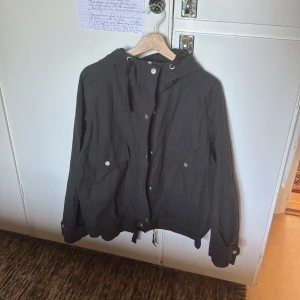 Svart vindjacka med huva och fickor - Svart vindjacka med huva och dragsko, stora fickor framtill med tryckknappar och justerbara ärmslut. Jackan har en enkel och clean look, perfekt för dig som gillar stilren och funktionell design. Materialet känns lätt och smidigt, och jackan har dragkedja och knappar framtill.