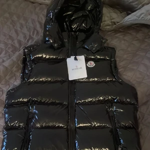 Svart dunväst från Moncler med huva - Snygg svart dunväst från Moncler med glansig finish och ikonisk logga på bröstet. Västen har dragkedja framtill, två fickor med dragkedja och en avtagbar huva. Perfekt för lager-på-lager och ger en riktigt clean streetstyle-vibe.