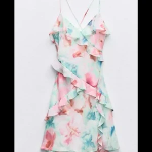 Pastellfärgad volangklänning med blommor - Superfin kort klänning med tunna axelband och volangdetaljer. Mönstret är blommigt i pastelliga färger som rosa, blått och grönt. Perfekt för sommarens alla tillfällen, lätt och luftig med romantisk vibe.