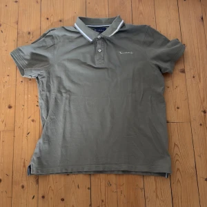 Olivgrön pikétröja från Bondelid - Snygg olivgrön pikétröja från Bondelid med klassisk krage och vita ränder. Tröjan har korta ärmar, två knappar framtill och broderad logga på bröstet. Perfekt för en clean och avslappnad stil.