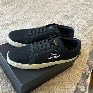 Svarta Saint Laurent sneakers - Svarta sneakers från Saint Laurent med vit sula och handskriven logga på sidan. Skorna har snörning, rund tå och är tillverkade i canvas och skinn. Guldtext på innersulan och klassisk low-top design. Perfekta för dig som gillar stilrena och exklusiva sneakers.