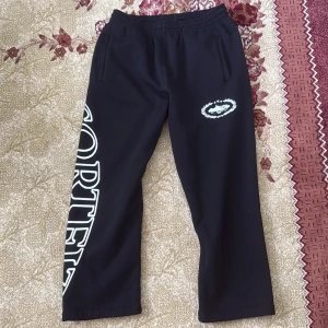 Svarta Corteiz mjukisbyxor med tryck - Svarta mjukisbyxor från Corteiz i storlek medium. Byxorna har resår i midjan, raka ben och stora vita tryck längs ena benet samt logga framtill. Perfekta för en chill och street-inspirerad look. Bra kvalite och väldig skön, knappt använd.