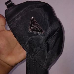 Svart keps från Prada  - Snygg svart keps från Prada med klassisk böjd skärm och justerbar rem bak. Kepsen är tillverkad i glansig nylon och har Pradas ikoniska trekantslogga i metall på sidan. Perfekt accessoar för en clean och lyxig streetstyle.