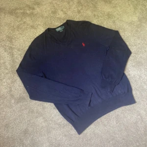 Mörkblå v-ringad tröja från Polo Ralph Lauren,strl M - Mörkblå v-ringad tröja från Polo Ralph Lauren i slim fit-modell. Tillverkad i 100% pima bomull och har det klassiska röda broderade logotypen på bröstet. Perfekt för en stilren och enkel look.