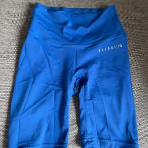 Blå träningstights shorts från Relode - Snygga blå träningsshorts från Relode med hög midja och tight passform i deras Mercy kollektion . Perfekta för träning eller gymmet, tillverkade i stretchigt syntetmaterial som sitter skönt och följer kroppen. Enkel design med diskret logga på ena benet.