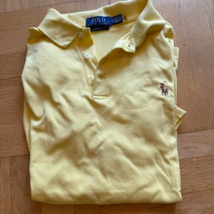 Gul pikétröja från Ralph Lauren - Snygg gul pikétröja från Ralph Lauren i bomull med klassisk krage, tre knappar och broderad logga på bröstet. Modellen är custom slim fit och har korta ärmar. Perfekt för dig som gillar stilren och sportig look.