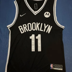 Brooklyn Nets Irving basketlinne Nike - Snyggt svart basketlinne från Nike med Brooklyn Nets-tryck och nummer 11 för Irving. Klassisk NBA-design med vita detaljer och loggor. Perfekt för dig som älskar basket och vill ha en sportig look. Materialet är lätt och andas, perfekt för träning eller sommaren.