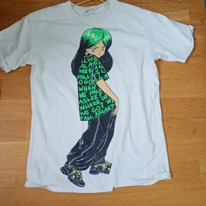 En fin billie elish tröja - Vit t-shirt från H&M med ett stort färgglatt tryck av en tecknad person med grönt hår och baggy byxor på framsidan. Text i grönt på motivet. Klassisk passform och rund hals. Perfekt för dig som gillar streetstyle och unika prints.