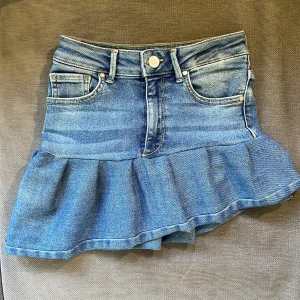 Blå jeanskjol med volang - Supersöt blå jeanskjol i kort modell med volang nertill. Klassisk femficksdesign. Perfekt för dig som gillar denim och vill ha en kjol med lite extra attityd.