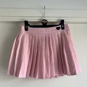 Rosa plisserad kjol från SHEIN - Supergullig rosa plisserad kortkjol från SHEIN i storlek S. Kjolen är i bomullsblandning och har en söt svart hjärt-detalj vid linningen. Perfekt för dig som vill ha en feminin och trendig look. Jag säljer även en vita. 