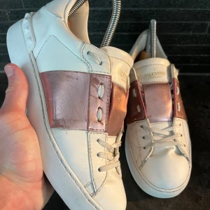 Valentino Garavani Open sneakers vit/rosa - Snygga Valentino Garavani Open sneakers i vitt skinn med metallicrosa band över vristen. Storlek 38, skick bra enbart någon repa. Tillbehör inget därav det lägre priset. NYPRIS 7’200kr