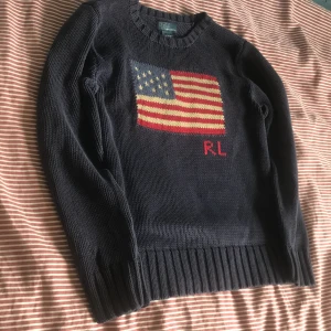 Mörkblå stickad tröja Polo Ralph Lauren - Mörkblå stickad tröja från Polo Ralph Lauren med amerikansk flagga och RL-broderi framtill. Tröjan har rund halsringning, ribbade muddar och är långärmad. Tröjan är lite mindre än storlek xs, så därmed xxs.