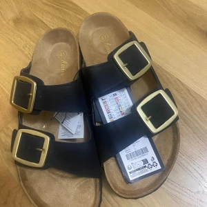 Svarta sandaler med stora spännen Amisu - Svarta sandaler från Amisu med två breda remmar och stora guldfärgade spännen. Fotbädden är i beige mockaimitation och sulan är svart med vågigt mönster undertill. Perfekta för varma dagar och enkel att matcha med allt.