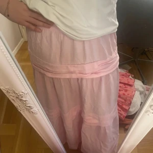 Ljusrosa långkjol i bomull - Superfin ljusrosa långkjol med volangdetaljer och skön, luftig känsla. Kjolen har en avslappnad passform och är gjord i ett tunt bomullstyg som passar perfekt till varmare dagar. Söt och romantisk stil som är lätt att matcha med olika toppar. Har även en kedja där bak. 