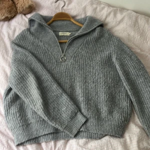 Grå stickad half zip hoodie från ONLY - Mysig grå stickad hoodie från ONLY i storlek S. Tröjan har en half zip-dragkedja med ringdetalj, huva och ribbade muddar. Perfekt för kalla dagar och enkel att styla med jeans eller leggings.