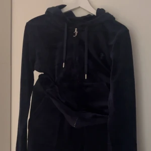 Mörkblått Juicy Couture set i velour - Mörkblått set från Juicy Couture i velour med dragkedja, huva och snörning. Logga på bröstet och snygga fickor med silverknappar framtill. Hoodie storlek M. Byxor storlek L. Väldigt fint skick! Båda delarna för 500 kr annars kan prisfisukteras vid köp av separat del! 