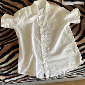 Vit kortärmad skjorta med ficka - Snygg vit skjorta med korta ärmar och klassisk krage. Skjortan har en bröstficka och knäppning framtill. Perfekt för en clean och fräsch look, lätt att matcha med jeans eller shorts.