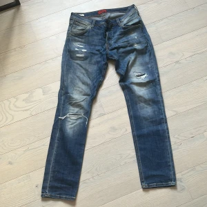 Jack & Jones Slim Glenn blå slitna jeans - Blå jeans från Slim Glenn med slitna detaljer och trasiga partier på benen. Jeansen har en slim passform och klassisk femficksdesign. Materialet är denim i bomull och färgen är mellanblå med ljusare tvättade partier. Skick 10/10!