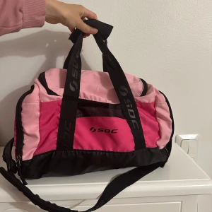 Rosa träningsväska från SOC - Säljer en snygg träningsväska från SOC i olika rosa nyanser med svarta detaljer och logga på handtagen. Väskan har både handtag och axelrem samt dragkedja upptill. Perfekt storlek för gymmet eller sportaktiviteter.
