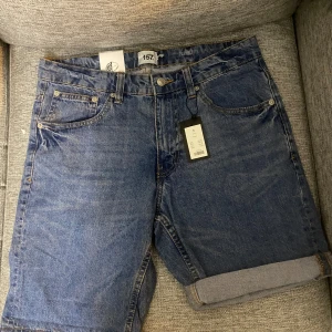 Blå jeansshorts från 157 - Klassiska blå jeansshorts från 157 med fem fickor, uppvikta benslut och normal passform. Shortsen har bälteshällor, knapp och dragkedja framtill samt snygga bakfickor. Perfekta för en chill sommardag.