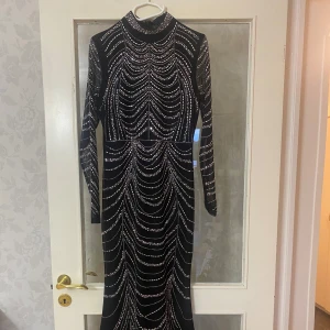 Svart glittrig långklänning från 3Q Fashion - Svart långklänning med glittriga stenar i ett mönster som påminner om pärlgardiner. Hög hals, långa ärmar i mesh och figurnära passform. Perfekt för dig som vill sticka ut med en riktigt glam look.