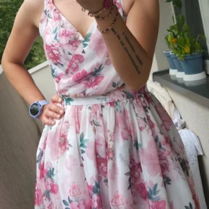 Blommig omlottklänning i rosa och vitt - Superfin kortklänning med omlottliv och v-ringning, täckt av blommigt mönster i rosa och grönt på vit botten. Klänningen har bred midja, dragkedja bak och är ärmlös. Perfekt för sommarens alla tillfällen!