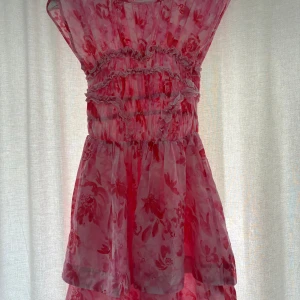Rosa blommig volangklänning - Supersöt kort klänning i rosa med blommigt mönster och volanger. Den har rynkade detaljer över byst och midja, samt ärmlös design med breda axelband. Köpt på bikbok, aldrig använd. Nypris 699kr