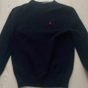 Mörkblå stickad tröja Polo Ralph Lauren - Mörkblå stickad tröja från Polo Ralph Lauren i storlek S. Klassisk modell med rund halsringning och röd broderad logga på bröstet. Tröjan har långa ärmar och ribbade muddar vid ärmslut och nederkant. Perfekt för lager-på-lager under kyliga dagar.