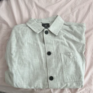 Ljusgrå overshirt från H&M - Snygg ljusgrå overshirt från H&M med klassisk krage och stora svarta knappar. Jackan har tre praktiska fickor framtill och är tillverkad i ett mjukt bomullsmaterial. Perfekt för lager-på-lager och enkel att styla till olika outfits.
