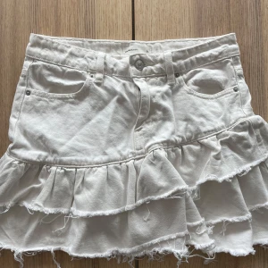 Vit jeanskjol med volanger - Supersöt vit jeanskjol från Perfect Jeans med volanger och råa kanter. Kjolen har klassiska fickor fram och bak, bälteshällor och knappstängning. Perfekt för dig som gillar en trendig och lekfull stil.