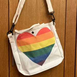 Pride väska regnbågshjärta canvas väska - 10 % av detta köp skänker jag till Karlskrona Pride  En canvasväska i lite mindre modell med ett stort härligt Pridehjärta på. Trycket är på båda sidor av väskan. Måtten är ca 23 x 24 cm. Den har handtag och dessutom en avtagbar axelrem som är justerbar. Knäpps med en knapp. Perfekt för sommaren. Helt ny.