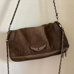 Brun Zadig & Voltaire väska  - Snygg brun axelväska i mocka med silverfärgad kedjerem och dragkedja. Väskan har en glittrig vingdetalj framtill som ger en cool touch. Perfekt storlek för det viktigaste och passar till många outfits.