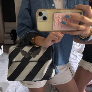 Randig handväska från MaxJenny! - Säljer en unik handväska från MaxJenny! i svart och silver med breda diagonala ränder. Väskan har ett coolt dödskalle-emblem med vingar framtill, kedjeremmar och handtag. Tillverkad i skinn med innerfack och dragkedja. Perfekt statement piece för dig som gillar edgy stil.