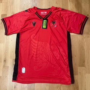 Albanien landslagströja 25/26 L - Albaniens fotbollströja från Macron i rött med svarta detaljer och emblem på bröstet. Tillverkad i lätt och ventilerande polyester.